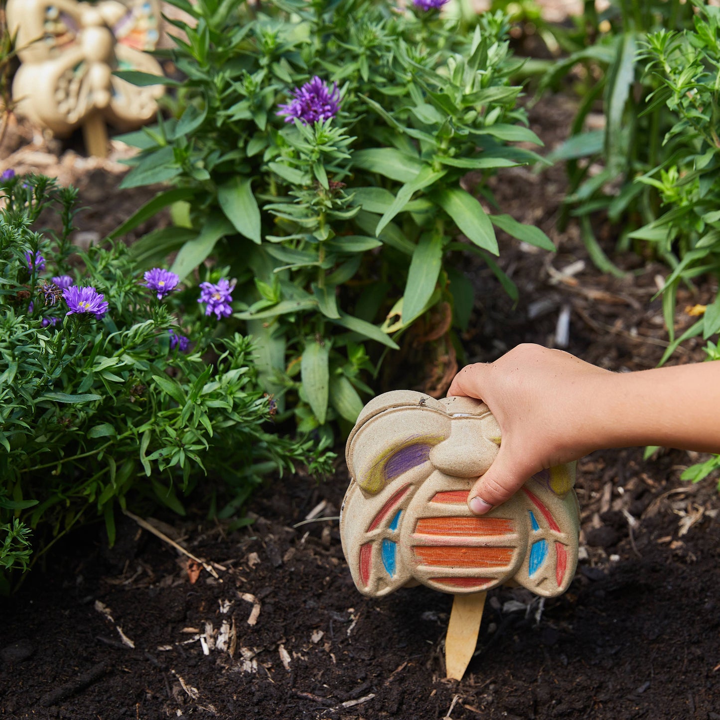 Hand holding a colorful butterfly shaped stake in a garden with plants and flowers.