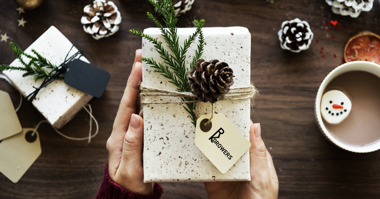 Hands holding a wrapped gift decorated with pine cones, greenery, and twine, with logo tag, above a rustic table with additional small gift boxes and pine cones