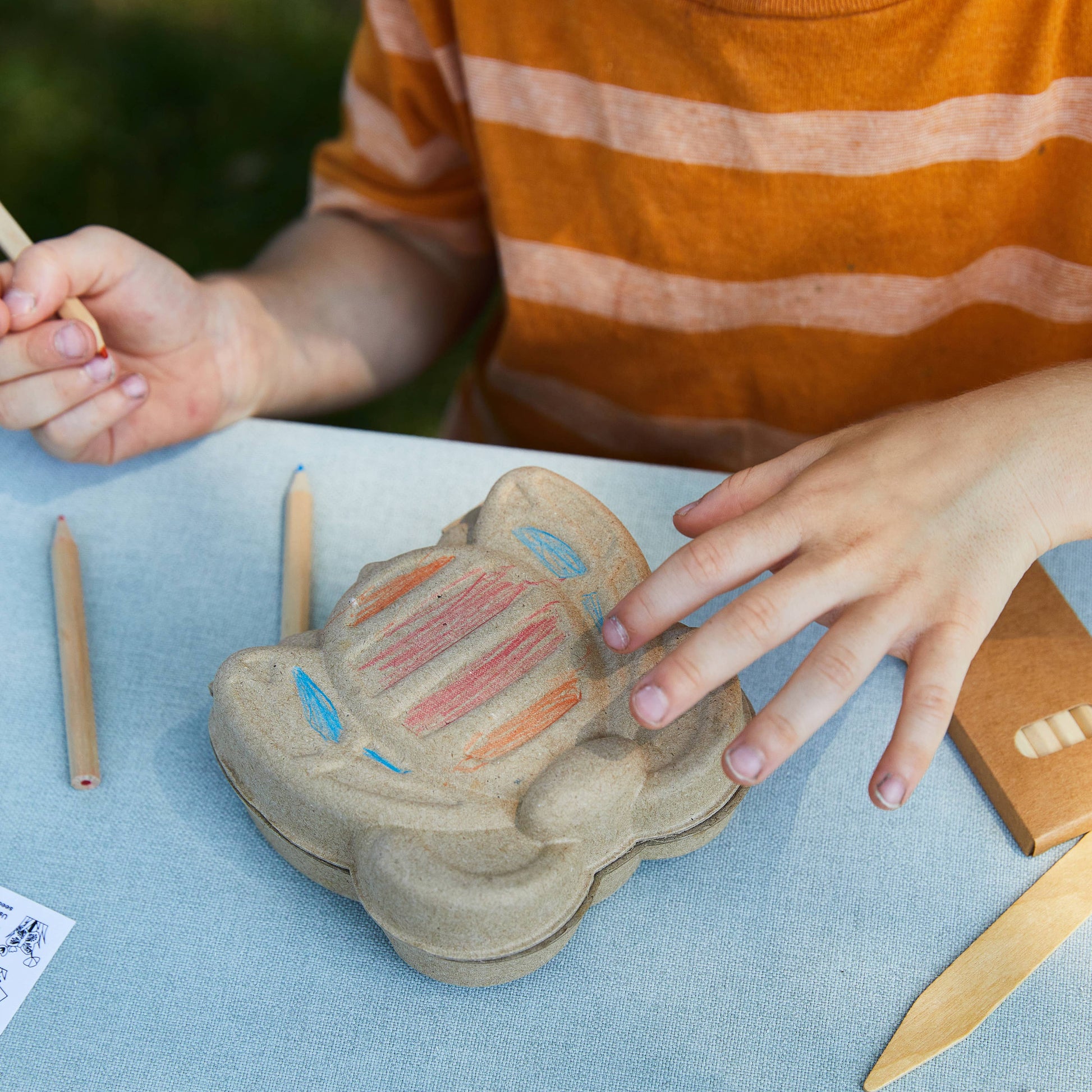 Child coloring a cardboard butterfly and wooden sticks on a blue surface