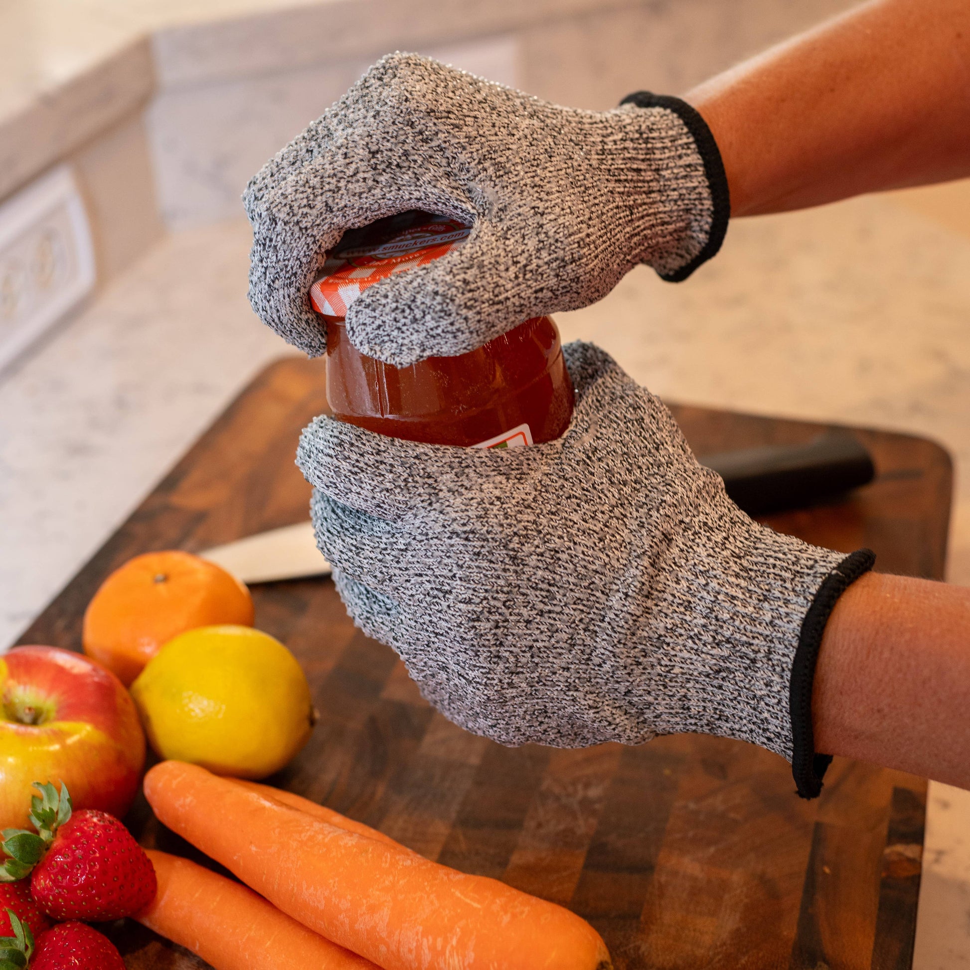 Person wearing cut-resistant gloves holding a jar on a kitchen counter with fruits and vegetables.